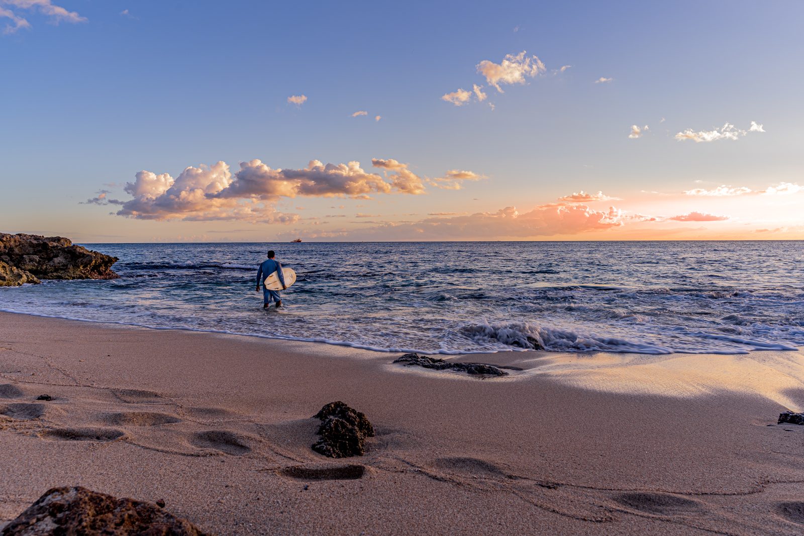 Surfer carrying a longboard into rich blue ocean light.